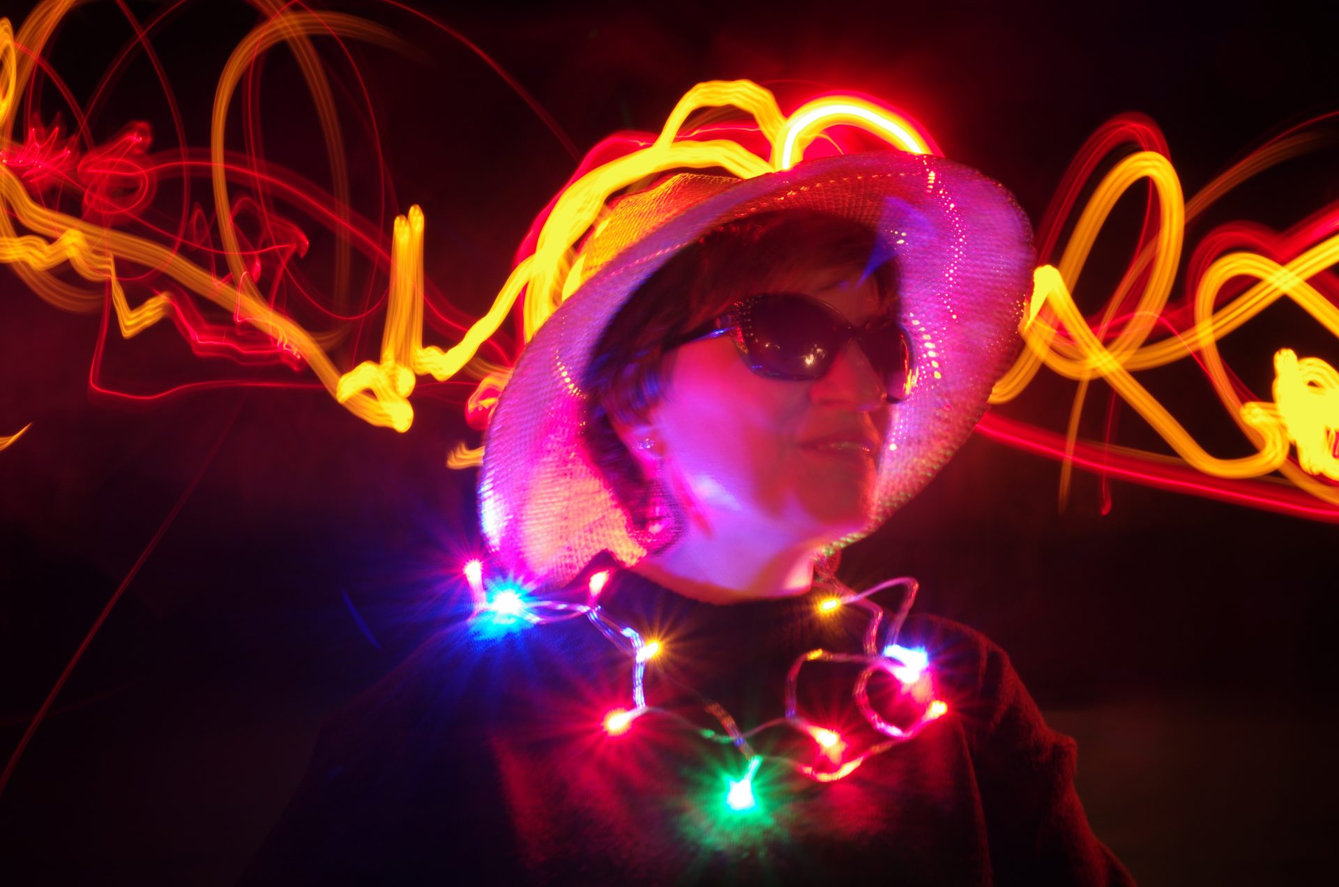 Frau mit Hut und Sonnenbrille in violettem Licht, um den Hals eine bunte Lichterkette, im Hintergrund ein gelbes Licht mit Bewegungsunschärfe.